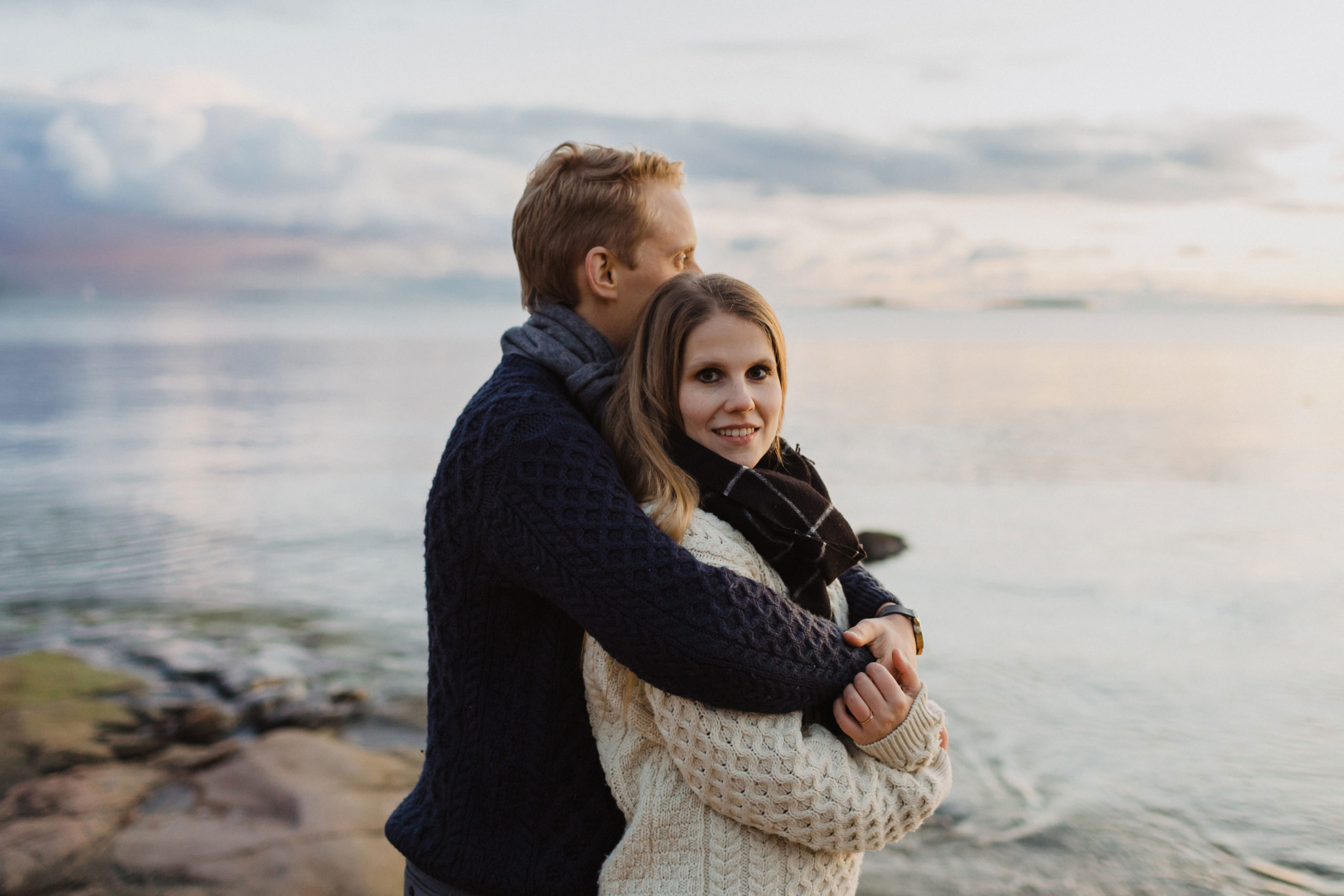 Man hugging his fiancé behind her back and she is looking straight to the camera smiling at the cliff at sunset in Lauttasaari in Helsinki